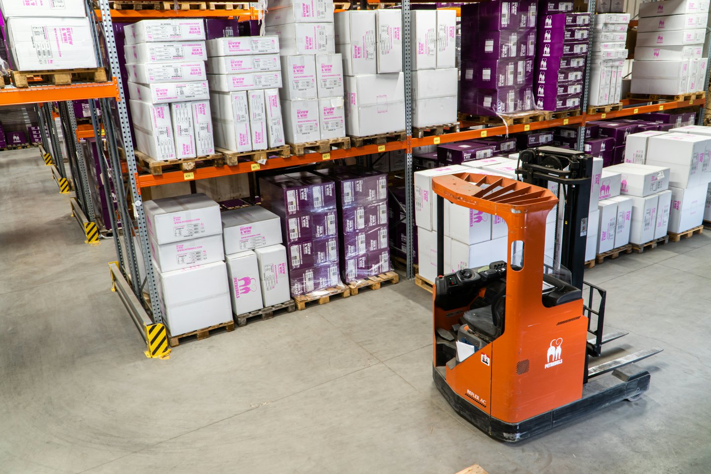 Orange gaffeltruck parkerad i en lagergång med staplade lådor.